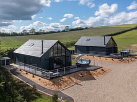 A modern farmhouse with two large glass-enclosed rooms, a hot tub, and a gravel driveway.