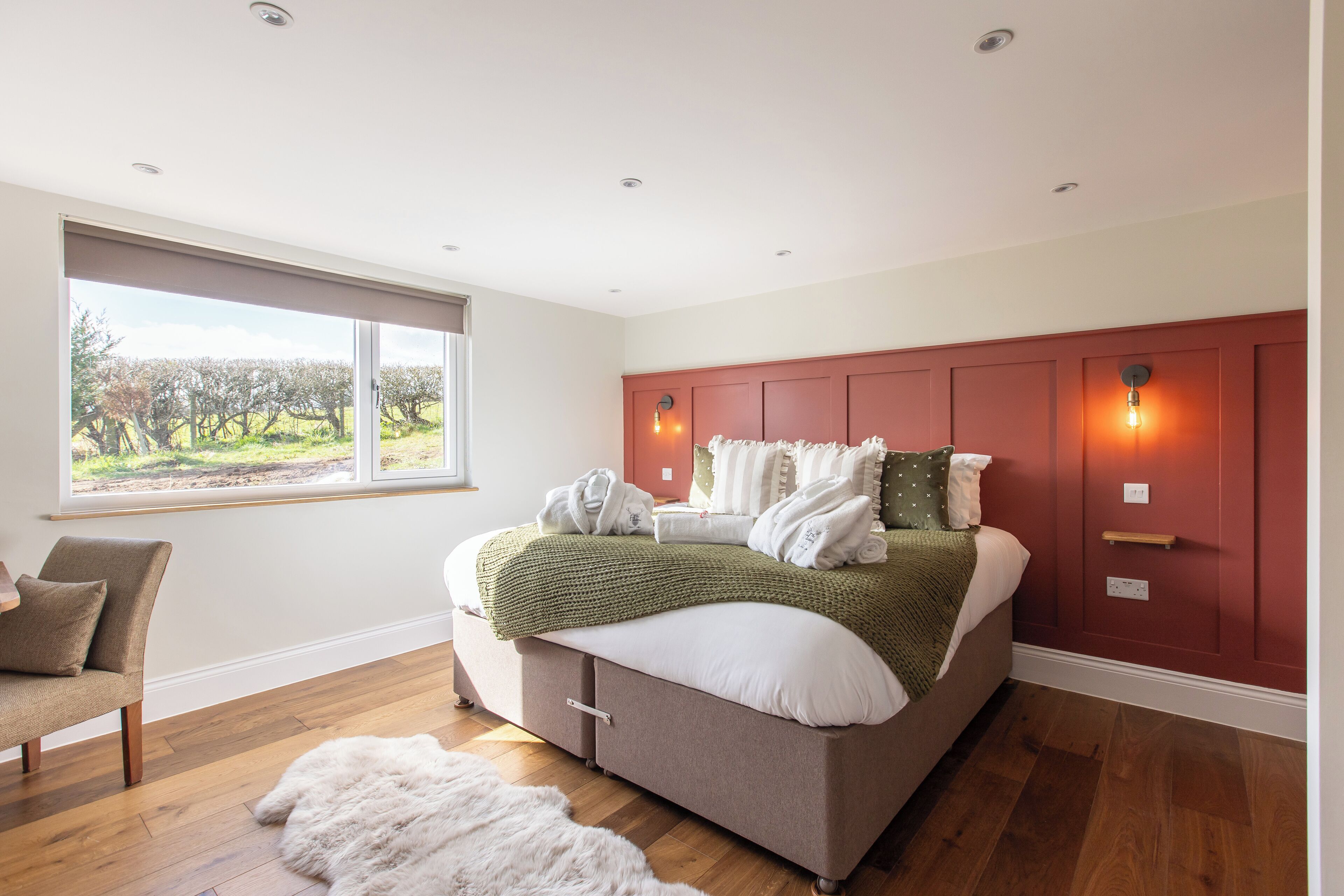 A bedroom with a large window, a bed with a green blanket, a chair, and a white fur rug.