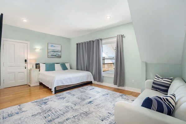 Une chambre à coucher avec un lit, une table de chevet, une fenêtre avec des rideaux, une vue sur l’extérieur et une porte blanche.