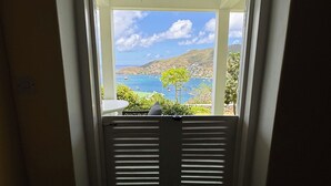 En vy genom ett fönster med fönsterluckor, med utsikt över ett kustlandskap med båtar och grönska.