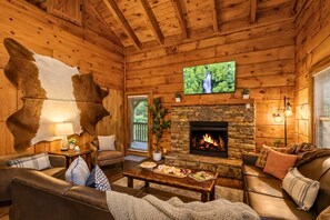 A cozy log cabin interior with a stone fireplace, leather sofas, and a television mounted on the wall.
