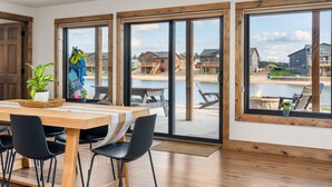 A dining area with a wooden table, black chairs, and a view of a lakeside community through large windows. - Lakefront Home w/Hot Tub & Beach – Sleeps 20 (New Lisbon)