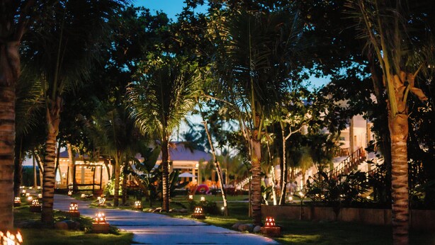 A pathway lined with palm trees and lit candles, leading to a building with a visible entrance.