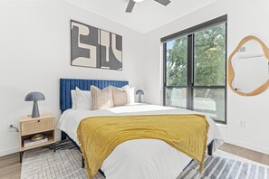 A modern bedroom with a large bed, a side table, a lamp, a bookshelf, and a large window with a view.