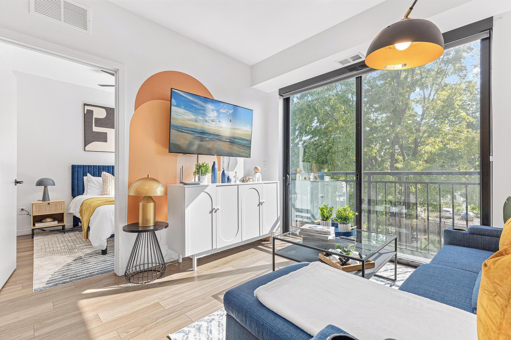 Ein modernes Wohnzimmer mit großem Fernseher, einer Couch, einem Couchtisch und einem Balkon mit Blick auf Bäume.