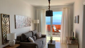 A living room with a sofa, a side table with a lamp, a decorative mirror, and a painting. There is a view of the ocean through a glass door. - Home2Book Esmeralda Beach, Resid. Playa Paraíso (Pajara)