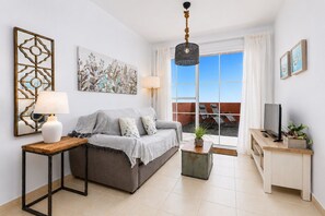 A living room with a sofa, a side table with a lamp, a television on a wooden stand, and a large window with a view of the outside. - Home2Book Esmeralda Beach, Resid. Playa Paraíso (Pajara)