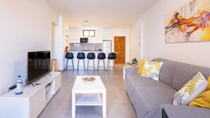 A modern living room with a grey sofa, a white coffee table, and a TV on a white stand. There is a kitchen in the background with a bar area. - Home2Book Sunset Terrace & Pool Haven (Puerto Santiago)