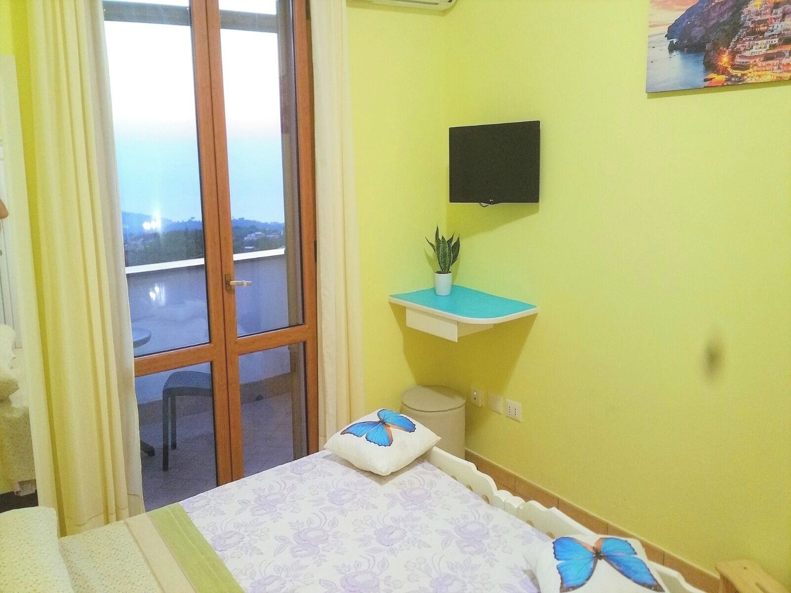 A bedroom with a bed, a small bedside table with a plant, a wall-mounted TV, and a window with a view of a balcony.