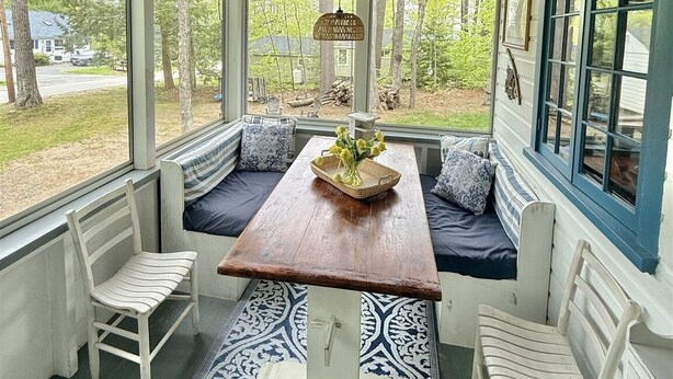 Une véranda couverte avec un plafond en bois, des murs blancs et une vue sur l’extérieur. On y trouve une table en bois avec des chaises et un banc avec des coussins.
