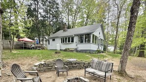 Un jardin avec un foyer en pierre, des fauteuils Adirondack en bois et une maison en arrière-plan.