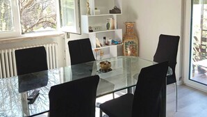 Un espace repas moderne avec une table en verre et des chaises noires, une étagère blanche agrémentée d’objets décoratifs, et une vue sur les arbres par la fenêtre.