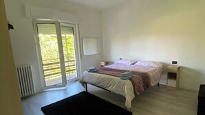 Une chambre avec un lit, un tapis noir, un plancher en bois, un radiateur et une porte-fenêtre coulissante donnant sur un balcon.