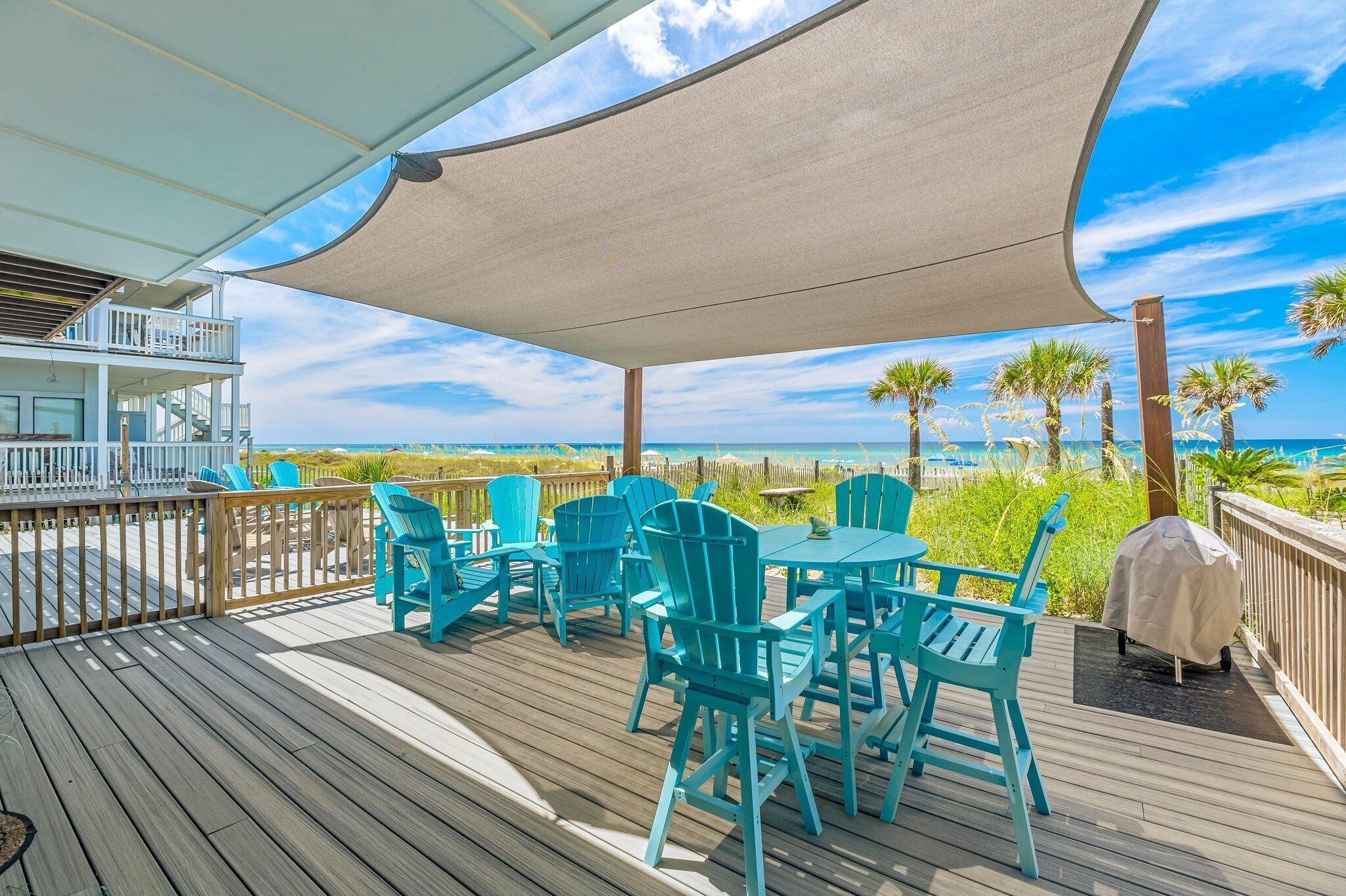 A wooden deck with turquoise chairs and a table, overlooking a beach with palm trees and the ocean.