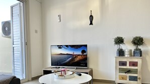 Una sala de estar con televisor sobre un mueble bajo, una mesa redonda de vidrio, un aparador con adornos y un cerramiento de vidrio corredizo.