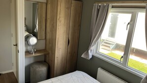 A bedroom with a bed, a wooden wardrobe, a vanity with a mirror, and a window with curtains. - Seaside Caravan in Eyemouth (Eyemouth)