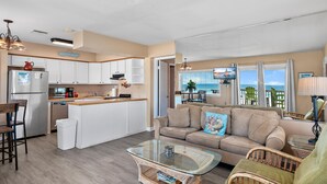 A modern kitchen with white cabinets, a dining area with a wooden table, and a living room with a beige sofa and wicker chairs. - Capri on the Gulf 108 (Destin)