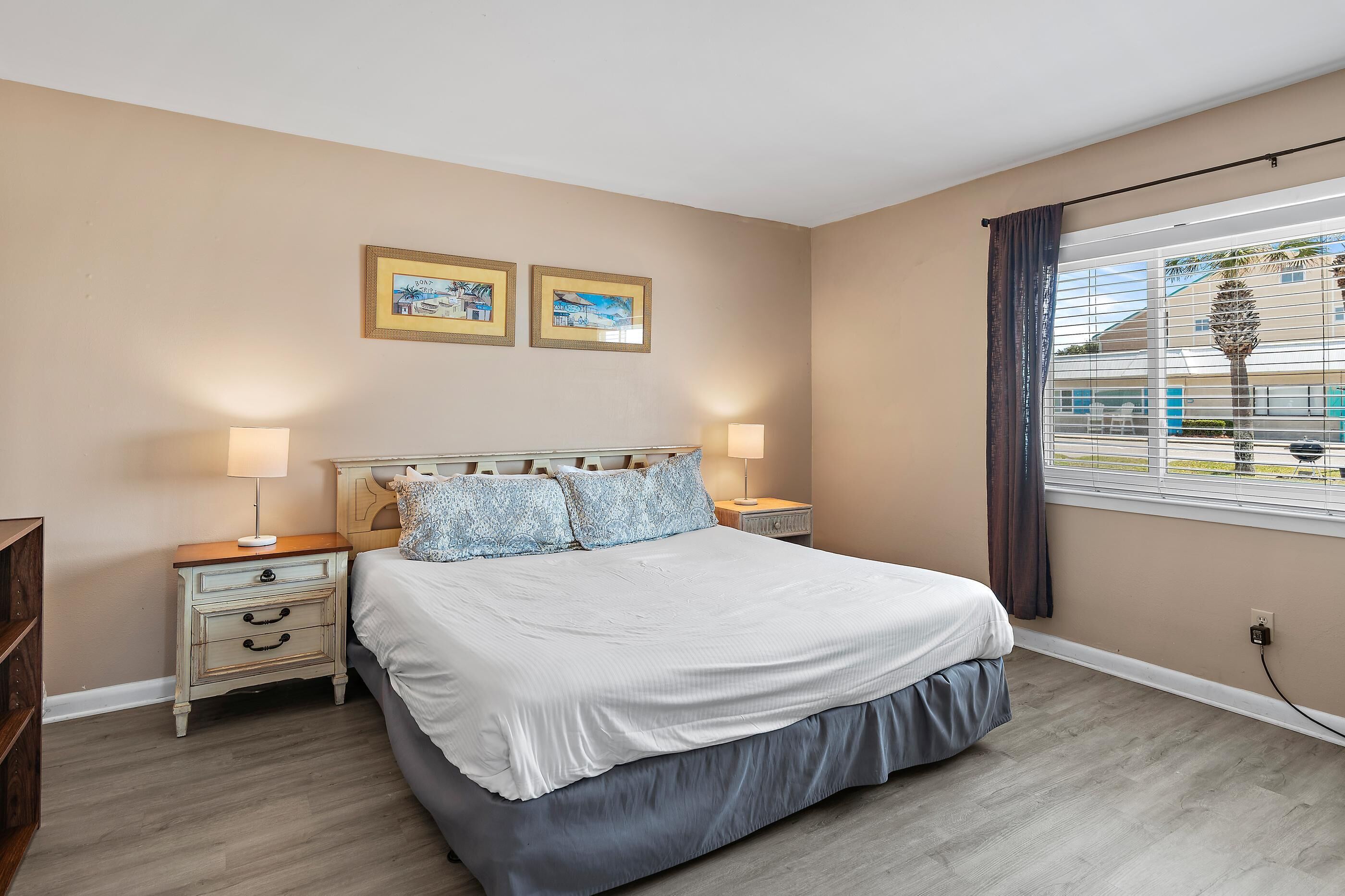 A bedroom with a bed, two bedside tables, a nightstand, a window with curtains, and two framed pictures on the wall.