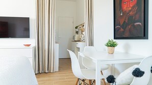 A modern hotel room with a flat-screen TV, a dining area with white chairs and a table, and a large painting on the wall. - Elegant Apt between sea, ponds and nature, 5 minutes from beaches, direct access to streetcar. (Pérols)