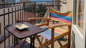 A balcony with a wooden chair and table, a striped towel, and a view of the cityscape and sea. - Sicilian Apt in Donnalucata-sleeps 4- 2 Min Beach (Scicli)