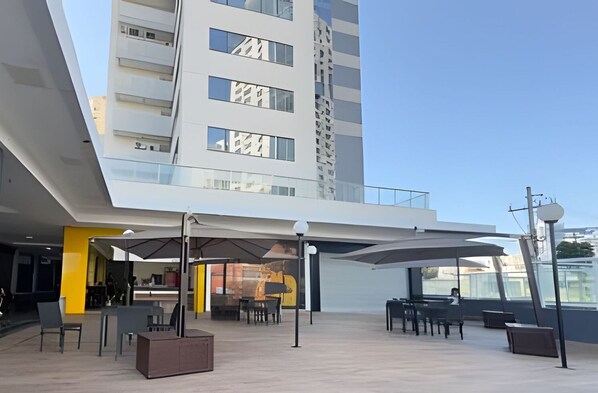 Uma área externa com mesas e cadeiras, parte coberta e um edifício moderno ao fundo.