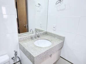 Casa de banho com lavatório de bancada em granito, duas torneiras, espelho e chuveiro.
