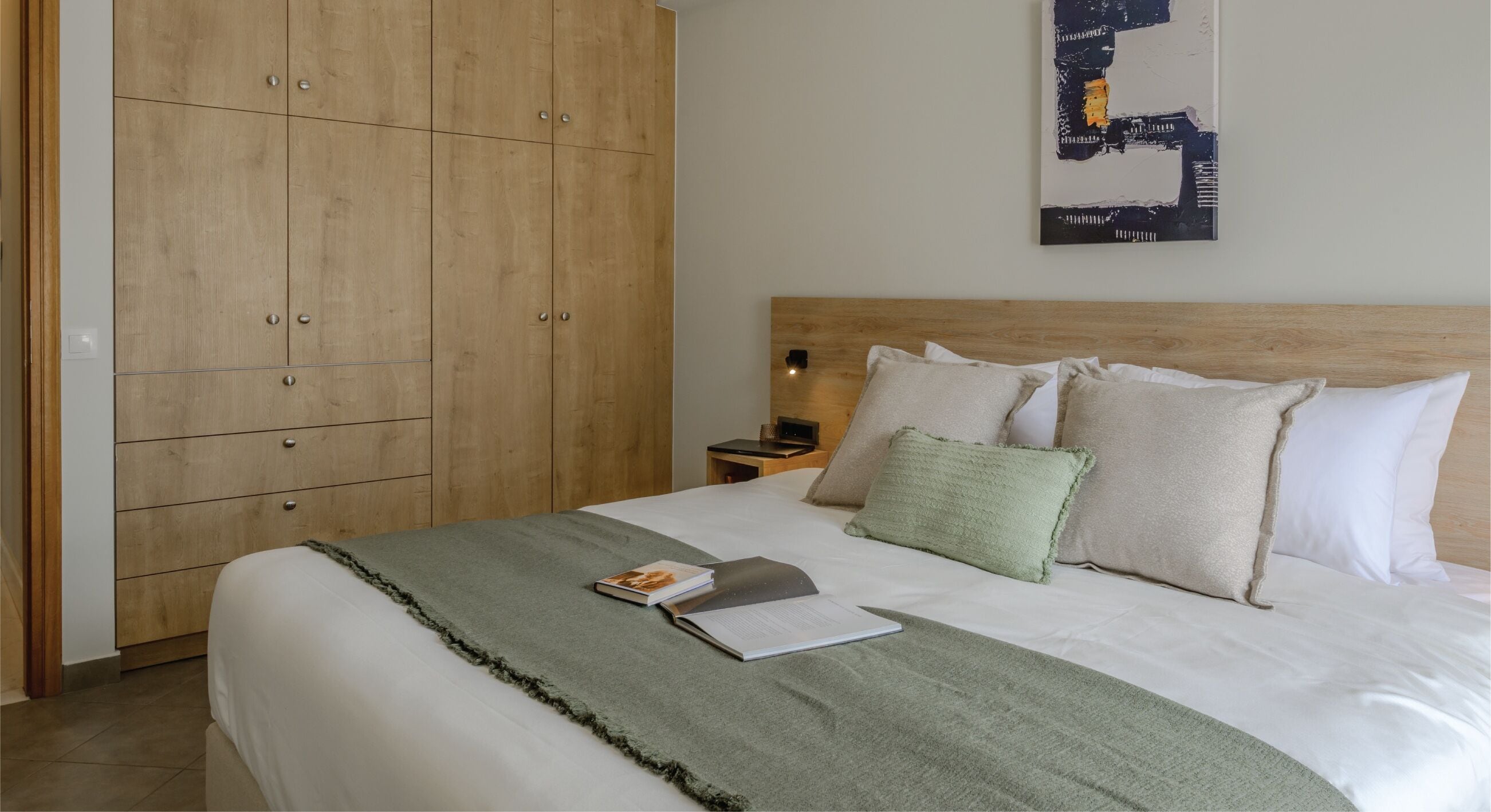 Una cama bien hecha con una manta verde, una mesita de noche con una lámpara y un armario de madera.