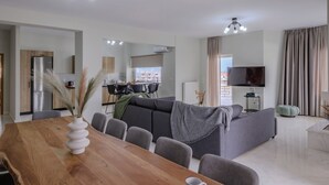 A modern living room with a grey sofa, a wooden dining table with chairs, a TV mounted on the wall, and a kitchen area in the background. - Jaax Apartment Airport by Airstay (Spata-Artemida)