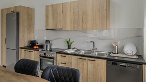 A modern kitchen with wooden cabinets, stainless steel appliances, a black countertop, and a dining area with a black upholstered chair. - Jaax Apartment Airport by Airstay (Spata-Artemida)