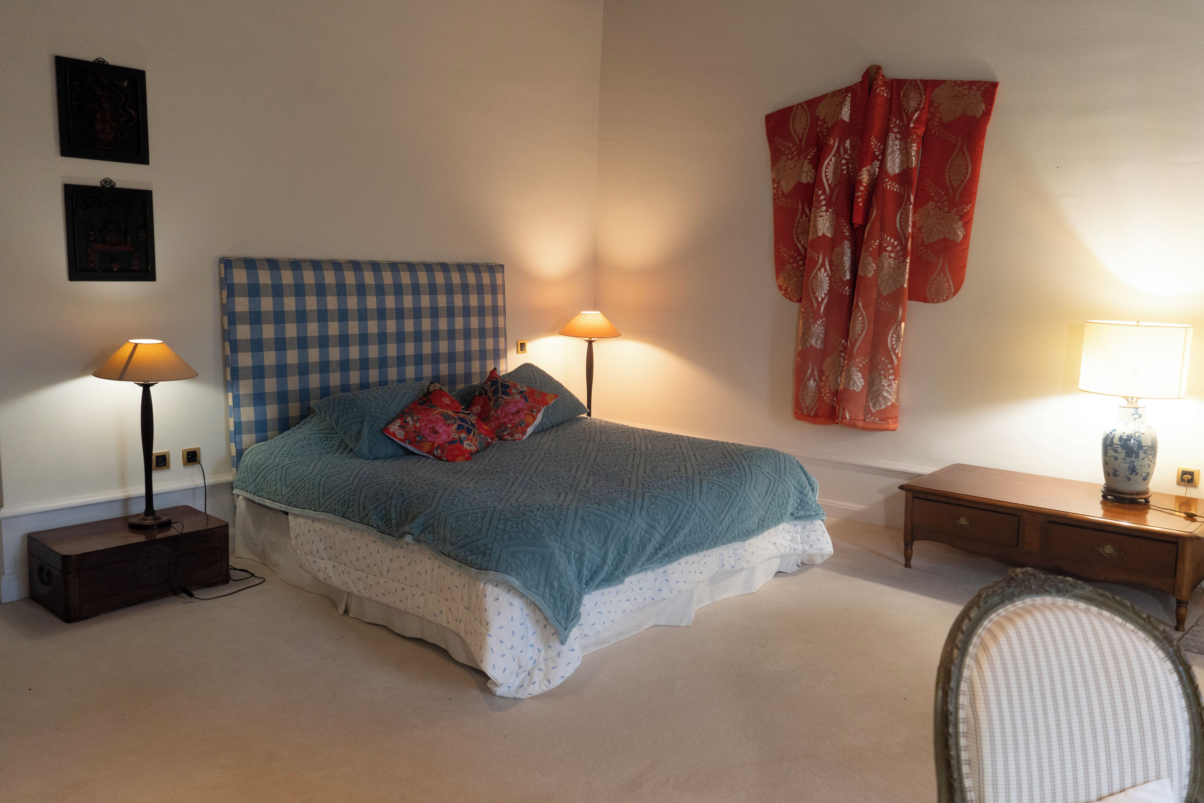 A bedroom with a checkered headboard, two bedside lamps, a bed with a patterned blanket, a red curtain, and a small bedside table with a vase.