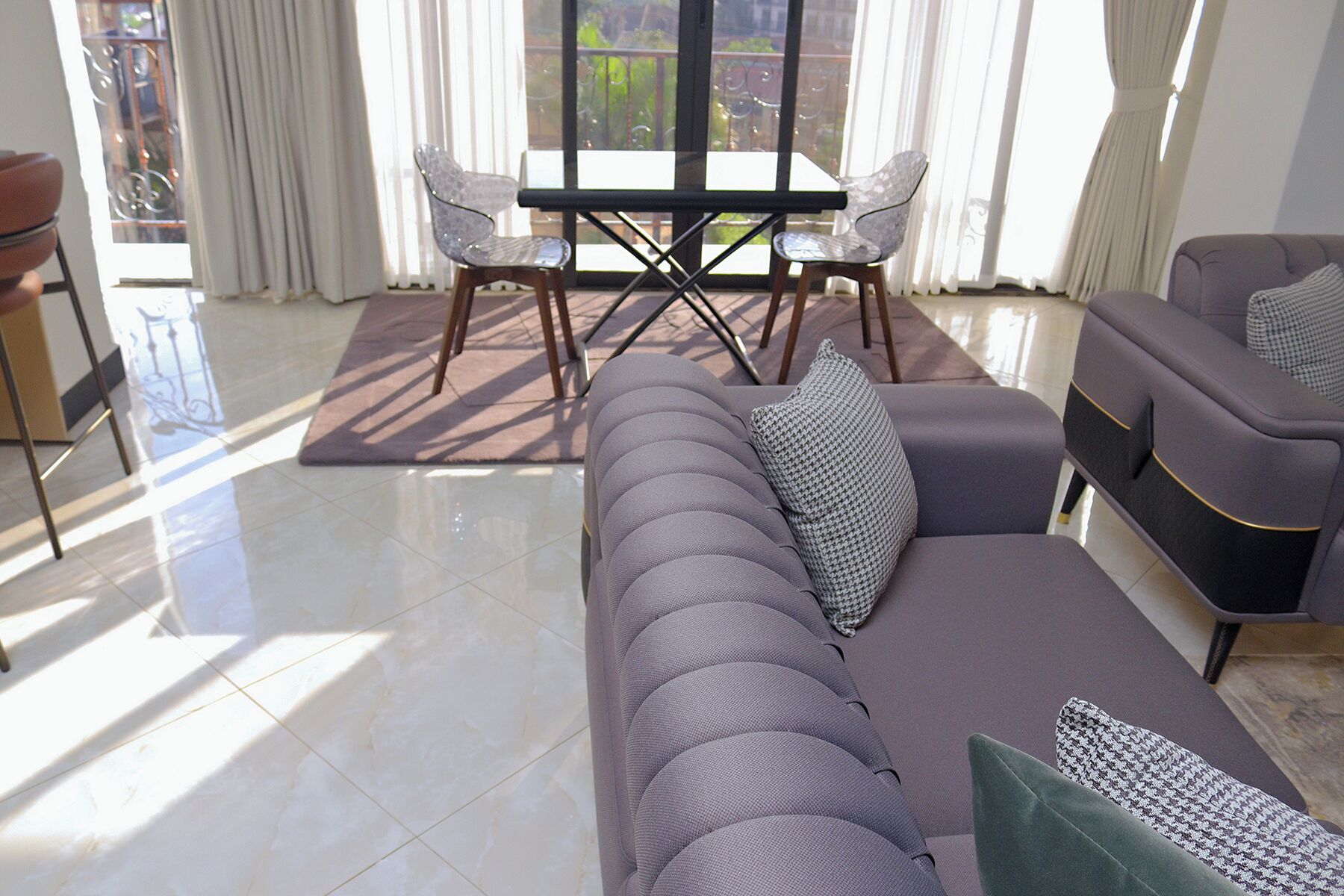 A modern living room with a grey sofa, a dining table with chairs, and large windows with curtains.