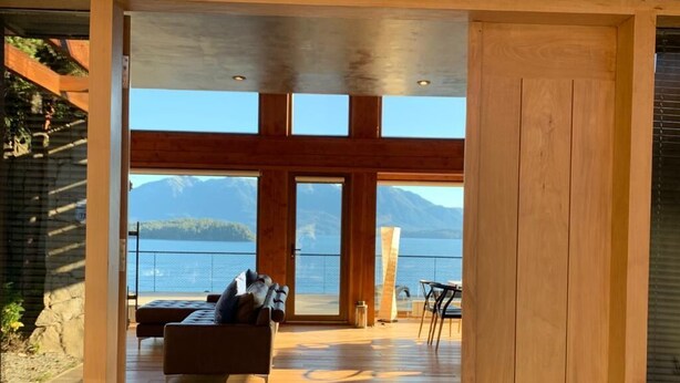 A spacious room with a wooden floor, a sofa, and a table. A large window offers a view of mountains and a body of water. - Tanière. Contemporary design house. Breathtaking lake view. (Villa La Angostura)