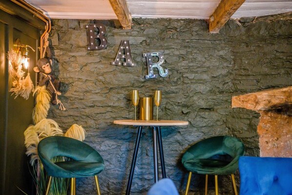 A rustic interior with a stone wall, a bar sign, two green chairs, and a small table with two golden glasses.
