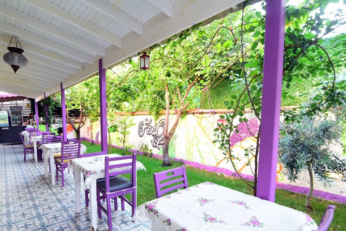 An outdoor cafe with purple chairs and tables, surrounded by greenery and a mural.
