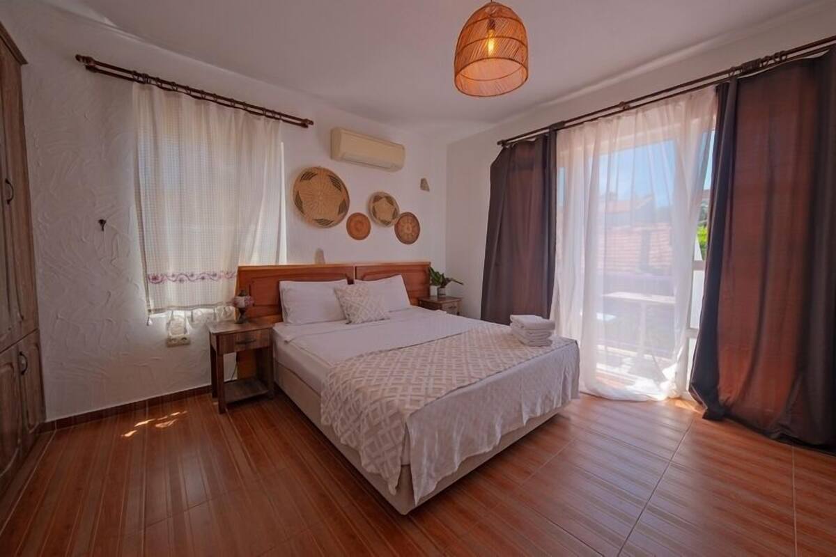 A bedroom with a wooden floor, a large bed with a white bedspread, a nightstand, a wall-mounted air conditioner, and a ceiling light fixture.