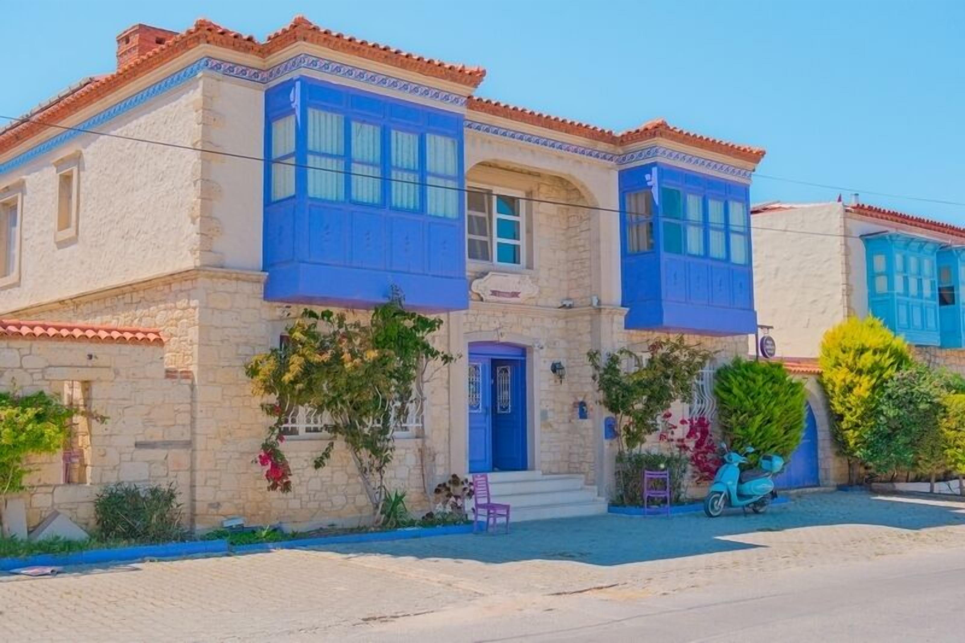 A two-story stone building with a blue door and windows, a scooter parked in front, and a bench outside.