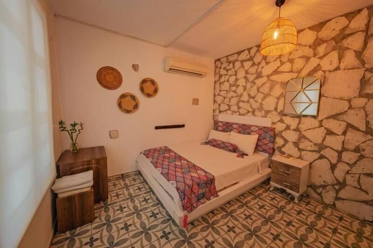 A bedroom with a bed, a nightstand, a wall-mounted mirror, and a stone wall.