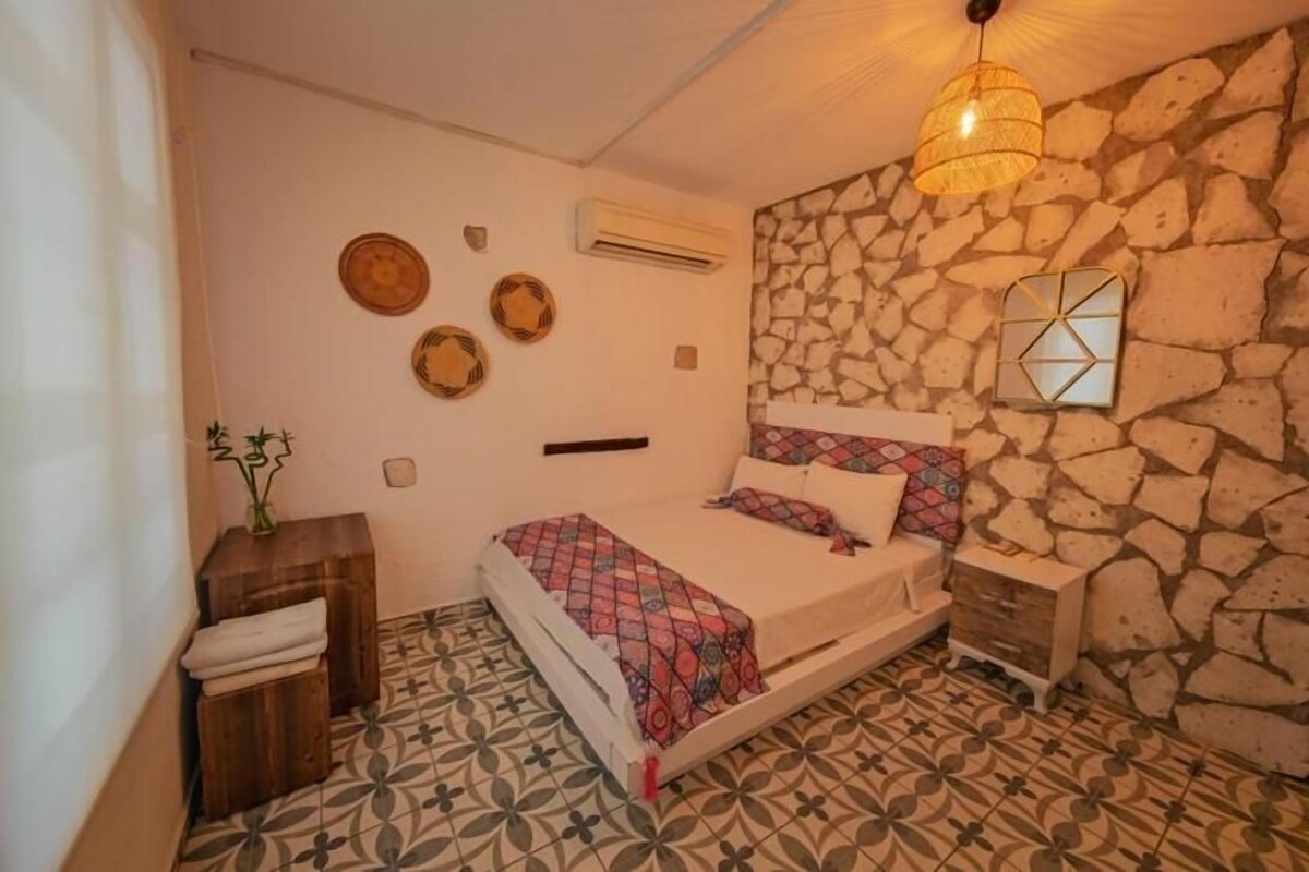 A bedroom with a bed, a nightstand, a wall-mounted mirror, and a stone wall.