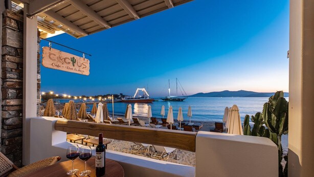 Eine Seeterrasse mit Blick auf das Meer, ein Boot und eine Brücke. Liegestühle und Sonnenschirme sorgen für Komfort, ein Tisch ist gedeckt mit einer Weinflasche und Gläsern.