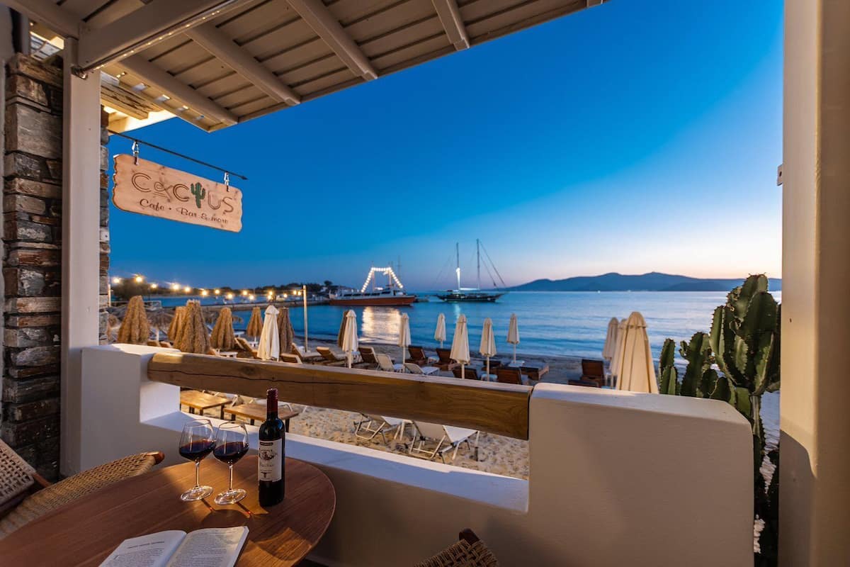 A seaside terrace with a view of the sea, a boat, and a bridge. There are lounge chairs and umbrellas, and a table with a bottle of wine and glasses.