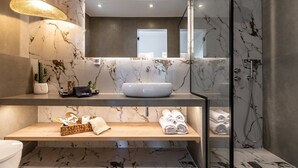 Ein modernes Badezimmer mit Marmor-Spritzschutz, einem großen Spiegel, einem weißen Waschbecken und einem Holzkosmetikschrank mit Handtüchern und Pflegeprodukten.