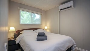 A neatly made bed with white bedding and folded towels, a bedside lamp, and a window with a view of trees. - Charming 4-bedroom house with AC, WiFi in brilliant Wasaga Beach (Wasaga Beach)