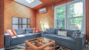 A cozy living room with a sloped ceiling, a sectional sofa with patterned cushions, a glass coffee table, and large windows offering a view of greenery. - Charming 4-bedroom house with AC, WiFi in brilliant Wasaga Beach (Wasaga Beach)