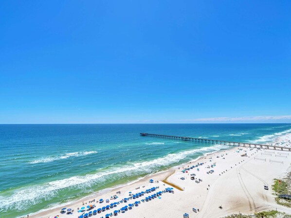 Ein Strand mit einer langen Pier, die sich ins Meer erstreckt, mehreren Reihen von Strandsonnenschirmen und einem Sandstrand.