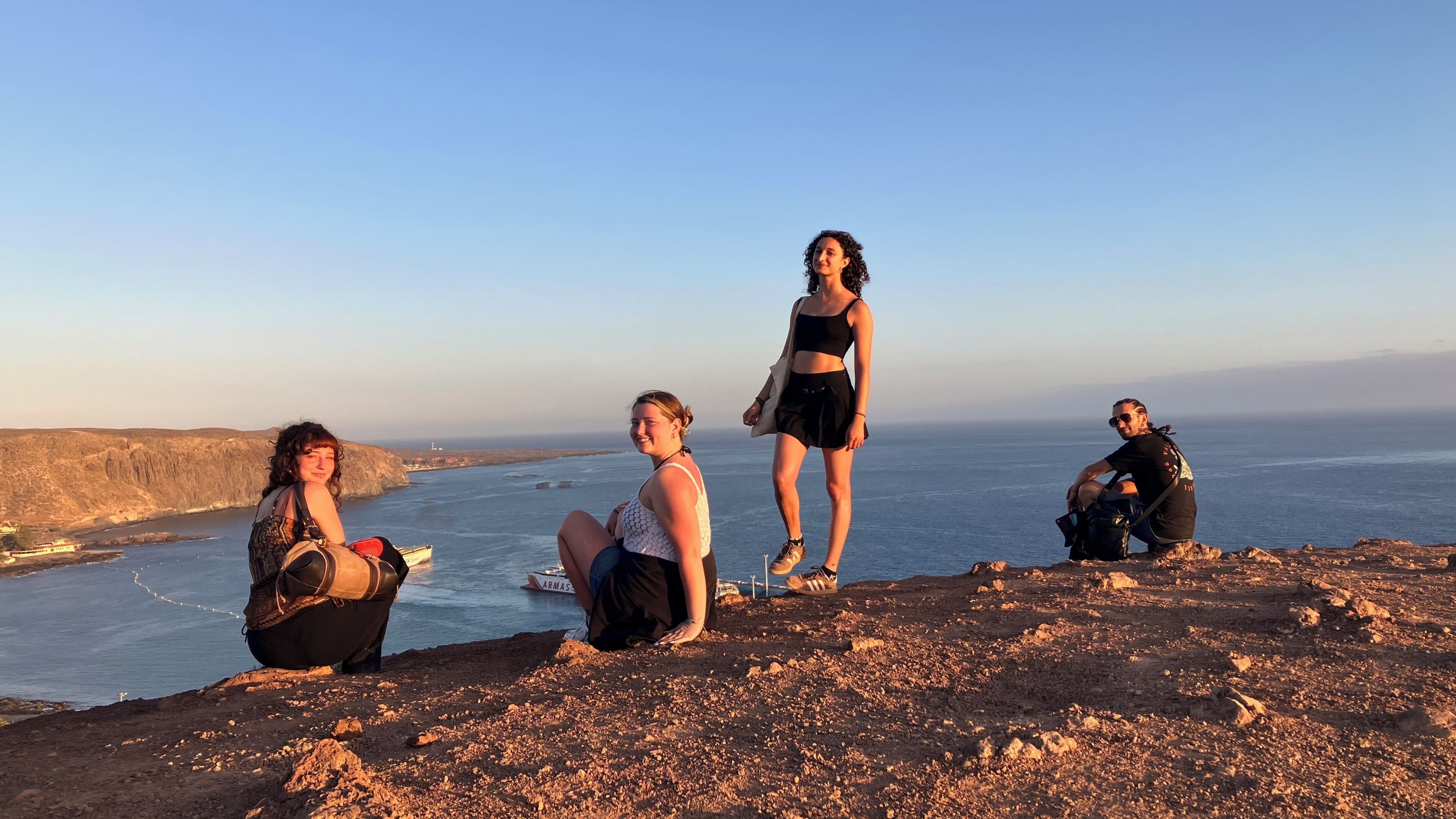 A group of people on a rocky cliff overlooking a coastal area with a clear view of the sea and distant land.
