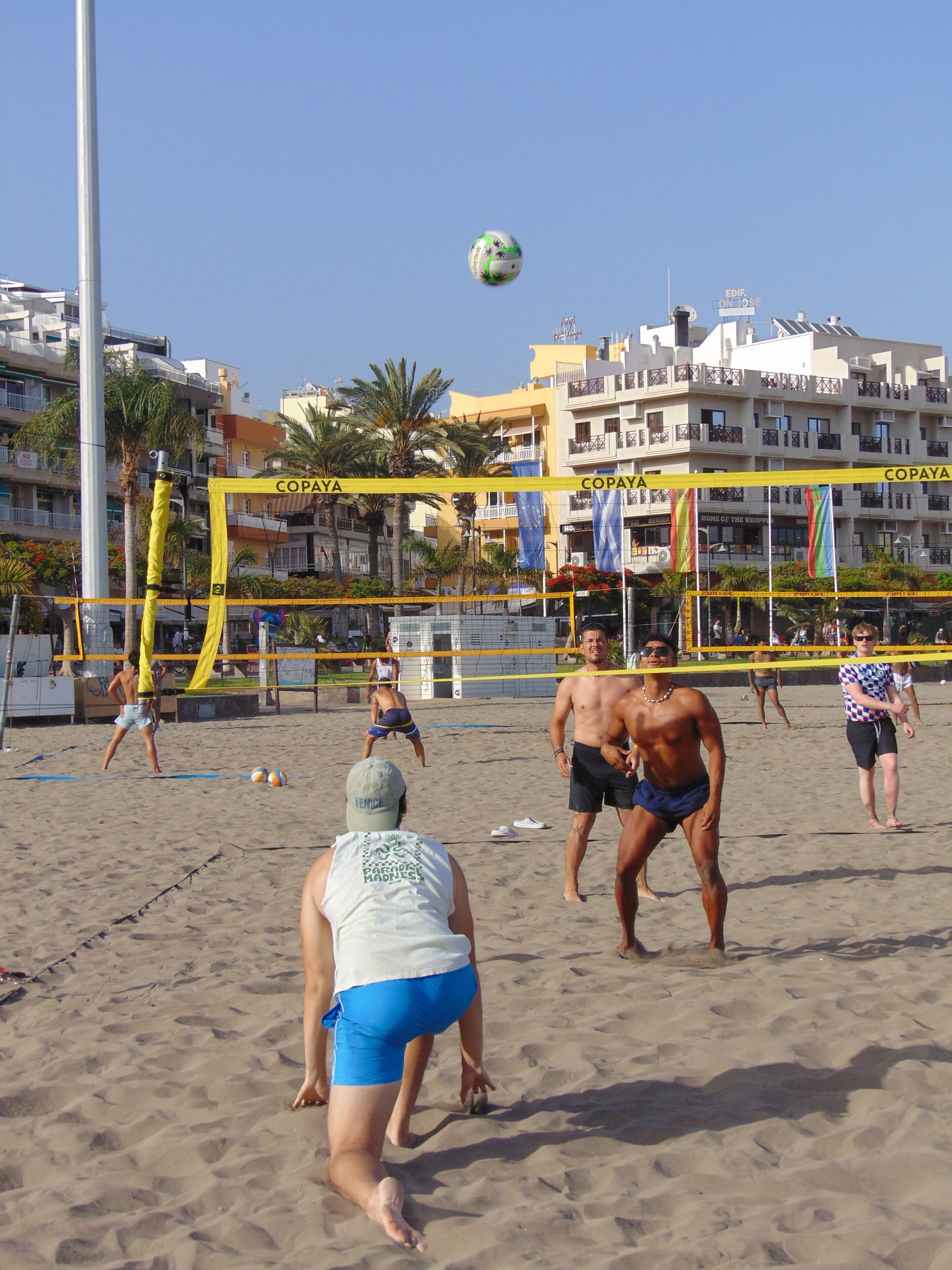 Ein Beachvolleyballspiel auf einem Sandplatz mit einem gelben Netz, umgeben von Gebäuden und Palmen.