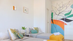 Habitación de hotel con dos camas, una mesita de noche, una lámpara y una puerta con un mural colorido.