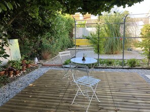 Um deck de madeira com mesa e cadeiras de metal, rodeado por plantas em vasos e uma área de jardim.