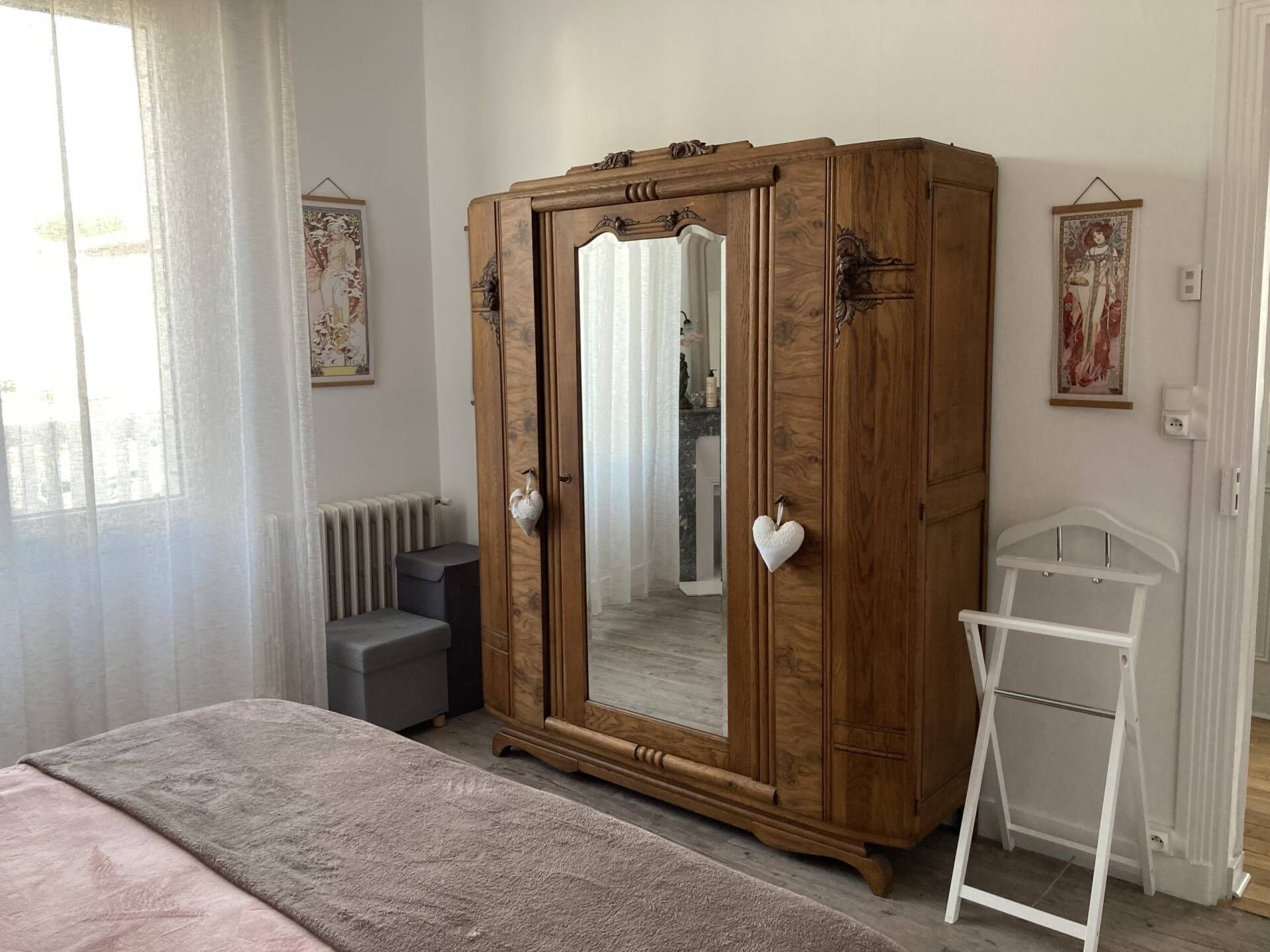 A bedroom with a large wooden wardrobe, a bed with a pink blanket, a white chair, and a radiator.