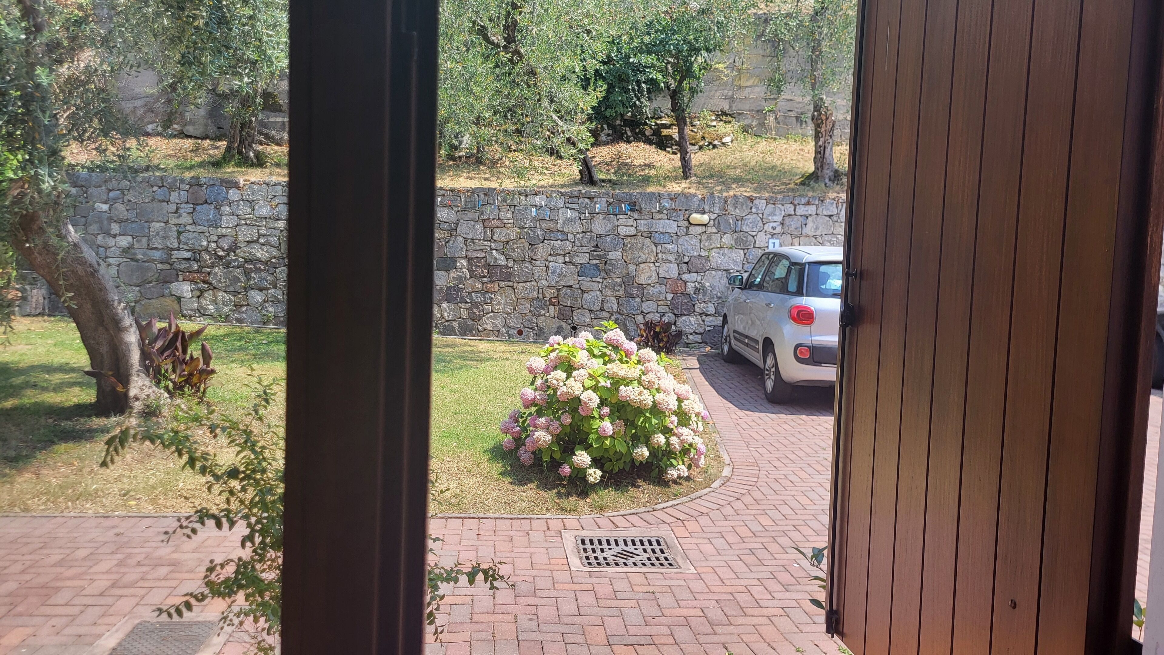 En stenmur, en bil parkerad på en gångstig av tegelstenar och en blomsterrabatt med rosa blommor.